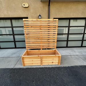 Puede incluir: Una jardinera rectangular de madera con un panel trasero de enrejado vertical integrado y alto. La estructura est&aacute; construida con tablones de madera natural de color marr&oacute;n claro con vetas visibles y un dise&ntilde;o de listones horizontales.