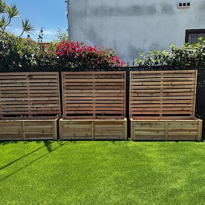 May include: Three wooden planter boxes with slatted privacy screens. The boxes are rectangular and made of light brown wood. The screens are composed of horizontal wooden slats. The planters sit on a green lawn, with a backdrop of greenery and a building.