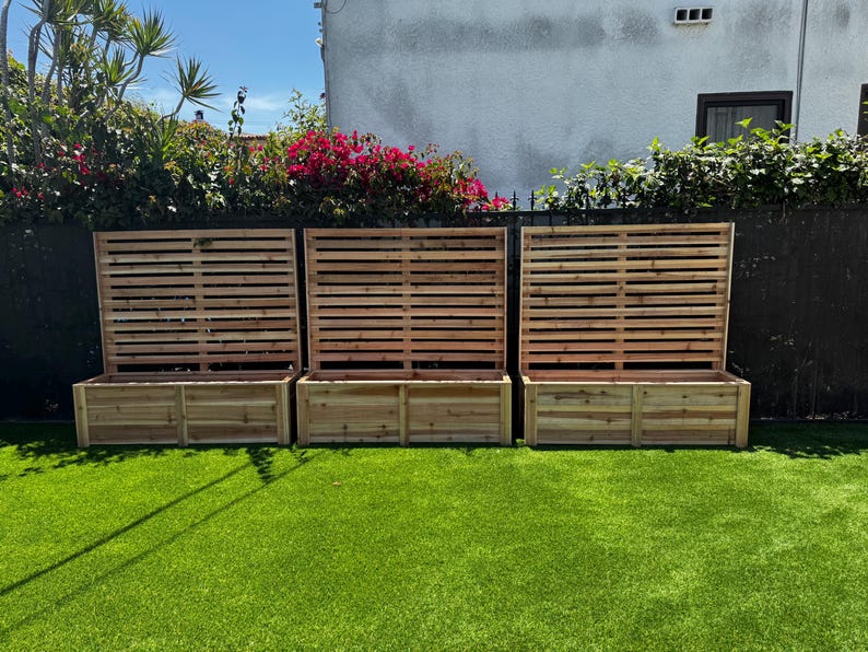 May include: Three wooden planter boxes with slatted privacy screens. The boxes are rectangular and made of light brown wood. The screens are composed of horizontal wooden slats. The planters sit on a green lawn, with a backdrop of greenery and a building.
