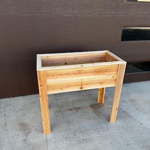 Puede incluir: Una jardinera elevada rectangular construida con madera de cedro de color marr&oacute;n claro. La jardinera cuenta con un recipiente profundo con un borde superior plano y est&aacute; sostenida por cuatro patas de madera verticales y resistentes.
