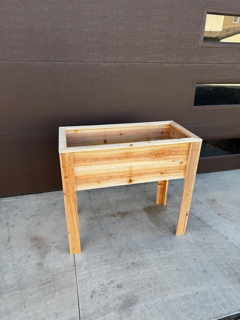 Puede incluir: Una jardinera elevada rectangular construida con madera de cedro de color marr&oacute;n claro. La jardinera cuenta con un recipiente profundo con un borde superior plano y est&aacute; sostenida por cuatro patas de madera verticales y resistentes.