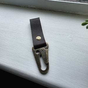 May include: A dark brown leather keychain strap featuring a brass-toned metal snap hook. The leather strip is folded and secured with a single brass-coloured metal rivet, connecting the strap to the spring-loaded carabiner-style metal clip.