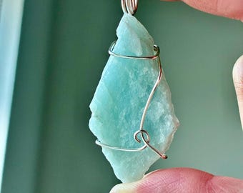 May include: A light blue, raw-cut gemstone pendant wrapped in silver-colored wire. The stone has a rough, natural texture and is suspended from a wire loop. The pendant is held up by a person's fingers.