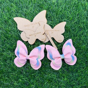 Puede incluir: Dos lazos para el pelo con forma de mariposa rosa con detalles iridiscentes y plantillas de mariposa de madera sobre un fondo verde. Las plantillas están etiquetadas como "BettyGirlyBows 3" y "#35".