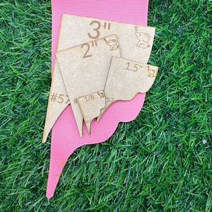 Puede incluir: Plantillas de madera para manualidades en varios tamaños, incluyendo 7,6 cm, 5,1 cm y 3,8 cm, con diseños de elefantes. Las plantillas están dispuestas sobre una forma rosa curvada, sobre un fondo de hierba verde.