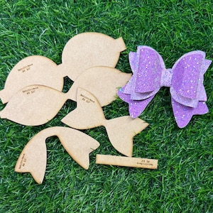 Puede incluir: Un conjunto de plantillas de lazo de madera y un lazo de purpurina morada terminado se disponen sobre una superficie verde. Las plantillas de madera tienen varias formas y tamaños, con el texto "BettyCuts Bow #3 3.5". El lazo terminado es en capas.