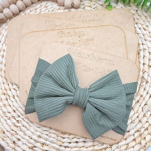 May include: A sage green ribbed bow on a wooden display card. The card has text that reads "Adult", "Toddler", "Baby", and "Newborn" with size ranges. The bow is centered on the card, with the text "Pretty Girly Bows" at the bottom.