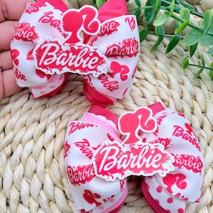 Puede incluir: Dos lazos para el pelo rosas y blancos con la palabra "Barbie" en escritura roja. Cada lazo presenta una silueta roja del perfil de Barbie. Los lazos están hechos de cinta y están colocados sobre un fondo tejido.