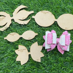 May include: A collection of wooden bow templates and a finished pink and white glitter bow are arranged on a green surface. The wooden templates are in various shapes and sizes, with text that reads "Betty Girl Bows 2.75" and the number "#27" on some.