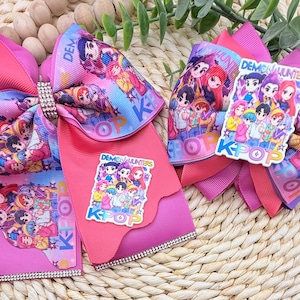 May include: Two pink hair bows with rhinestone accents. One bow features a large bow with a center rhinestone piece and a printed design with the words "Demon Hunters" and "K-Pop". The other bow has a sticker with the same design.