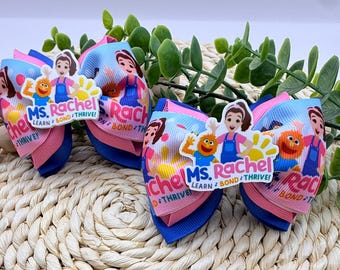 May include: Two Ms. Rachel hair bows. Each bow features a colorful design with the text "Ms. Rachel Learn Bond Thrive!" and illustrations of characters. The bows have pink, blue, and white ribbons.