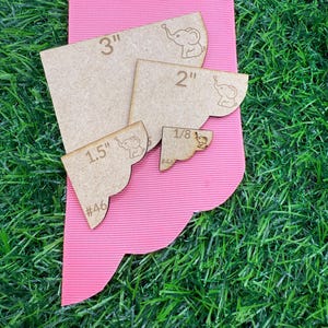 May include: A collection of wooden craft templates in various sizes, ranging from 1/8 inch to 3 inches. Each template features an elephant design and measurement markings. The templates are arranged on a pink ribbon against a backdrop of green grass.