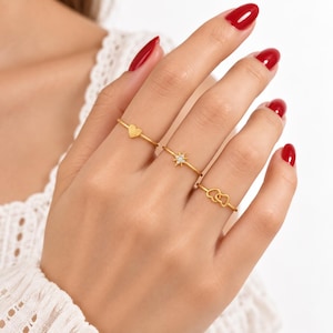 May include: Three thin gold rings worn on the fingers. The first ring features a small heart charm, the second ring displays a starburst design with tiny embedded stones, and the third ring consists of two interlocking heart shapes in a gold finish.