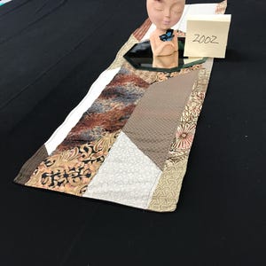 May include: A rectangular fabric table runner featuring a patchwork design of various brown, tan, and cream patterns. A beige head-shaped planter with a blue butterfly accent sits on a small octagonal mirror, next to a note with the text 2002.