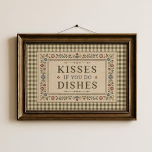Op de afbeelding: Een ingelijste kruissteek met een groen-wit geblokte achtergrond. De centrale tekst luidt "KISSES IF YOU DO DISHES" in donkergroene letters, omringd door een decoratieve rand van kleine rode, blauwe en gele bloemmotieven.