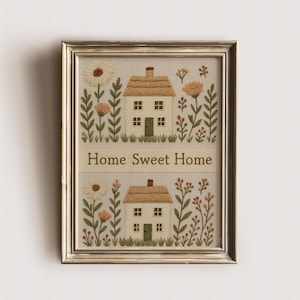 Puede incluir: Arte bordado enmarcado que presenta dos pequeñas casas con tejados de paja, rodeadas de flores y vegetación bordadas. La frase "Home Sweet Home" está cosida en el centro. El marco es de color dorado metálico.