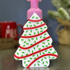 Op de afbeelding: Een roze zeepdispenser met een kerstboomvormige hoes. De witte boom heeft rode strepen en groene hagelslag. De dispenser heeft een roze pomp en staat op een reflecterend oppervlak.