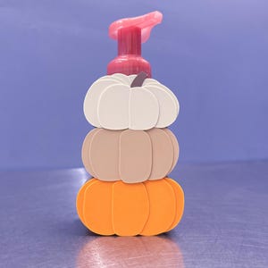 May include: A soap dispenser featuring a stack of three pumpkins in cream, beige, and orange colors. The dispenser has a dark red pump mechanism at the top. The pumpkins are arranged vertically, with a small dark brown stem on the top cream pumpkin.