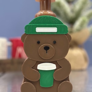 May include: A brown bear-shaped soap dispenser with a green beanie and a green coffee cup. The dispenser has a brown pump and is designed to resemble a friendly bear holding a coffee. The bear's eyes and nose are black.