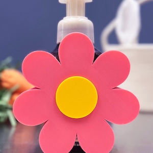 Peut inclure: Un couvercle de distributeur de savon en forme de fleur rose avec un centre jaune. Le couvercle est placé sur un distributeur de savon de couleur foncée avec une pompe blanche. L'arrière-plan est bleu flou.