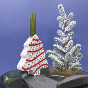 May include: A white Christmas tree-shaped straw topper with red and green accents sits atop a dark gray lid. A green straw extends from the topper. A small, snow-covered artificial tree is in the background against a blue backdrop.