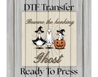 May include: DTF Transfer featuring a Halloween design with three cartoon geese. One goose is dressed as a ghost, and two are wearing witch hats. The text reads "Beware the honking Ghost" and "Ready To Press". The background is a wood grain pattern.