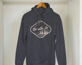 May include: Dark gray hoodie with a front pocket and drawstring hood. The hoodie features a cream-colored diamond-shaped graphic with the words "Breathe In" and a landscape design of trees, a road, and clouds.