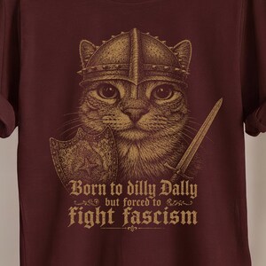 Könnte beinhalten: Burgunderfarbenes T-Shirt mit einer Katze, die einen Helm trägt und ein Schwert und einen Schild hält. Die Grafik enthält den Text "Born to dilly Dally but forced to fight fascism."