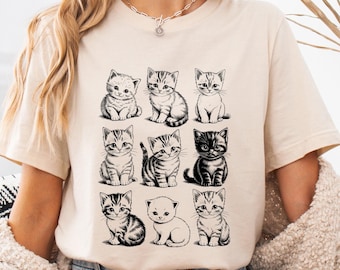 May include: A cream-colored t-shirt featuring a grid of nine adorable kitten illustrations in black ink. The kittens are depicted in various poses, with detailed fur patterns. The shirt is part of a casual wear collection.