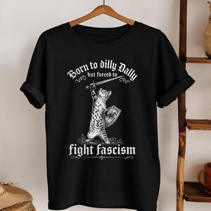 Könnte beinhalten: Schwarzes T-Shirt mit einer Grafik einer Katze, die ein Schwert und einen Schild hält. Der Text lautet "Born to dilly Dally but forced to fight fascism" in gotischer Schrift. Das Shirt hängt an einem Holzkleiderbügel.