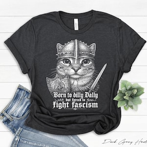 Könnte beinhalten: Dunkelgraues meliertes T-Shirt mit einer detaillierten Illustration einer Katze mit Helm, Schwert und Schild. Der Text lautet: "Born to dilly Dally but forced to fight fascism."