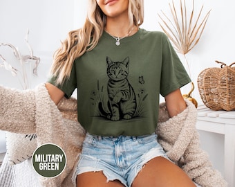 May include: Military green t-shirt featuring a black and white illustration of a cat with a butterfly and plants. The shirt is a relaxed fit. The text "MILITARY GREEN" is in a circular label in the bottom left corner.