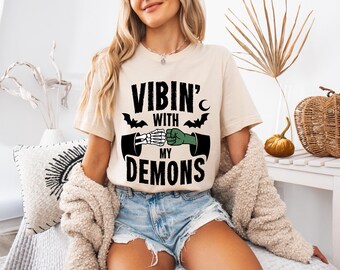 Puede incluir: Camiseta beige con el texto negro "Vibin' With My Demons". El diseño presenta una mano de esqueleto chocando los puños con una mano enguantada verde, con murciélagos y una luna creciente. La camiseta es parte de una colección temática de Halloween.