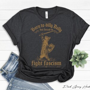 Könnte beinhalten: Dunkelgraues meliertes T-Shirt mit einer Illustration einer Katze, die ein Schwert und einen Schild hält. Der Text lautet "Born to dilly dally but forced to fight fascism" in einer Vintage-Schriftart. Das Shirt wird mit Jeans kombiniert.