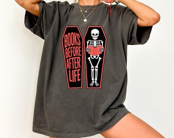 Puede incluir: Camiseta gris oscuro con el texto "BOOKS BEFORE AFTER LIFE" y un dibujo de un esqueleto leyendo un libro en un ataúd. El diseño es rojo y blanco.