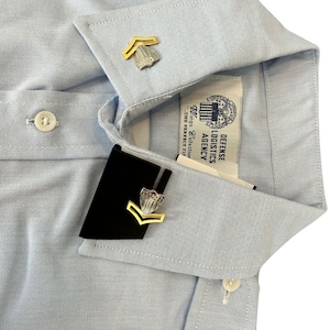 Puede incluir: Camisa azul claro con insignias doradas y plateadas en el cuello. La camisa tiene una botonadura frontal y una etiqueta que dice "Defense Logistics Agency". La camisa está hecha de una tela lisa y tejida.