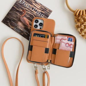 Puede incluir: Una funda de teléfono de cuero marrón claro con diseño de cartera, con cierre de cremallera y múltiples ranuras para tarjetas. La funda está abierta, revelando tarjetas y efectivo. Se adjunta una correa a juego. Una revista con el texto "Day: Plan" está en el fondo.