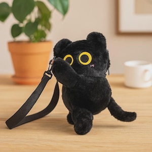 Może przedstawiać: Czarna pluszowa torba w kształcie kota z dużymi żółtymi oczami i czarnym paskiem. Kot ma puszystą fakturę i mały różowy rumieniec na policzkach. Torba stoi na drewnianej powierzchni.