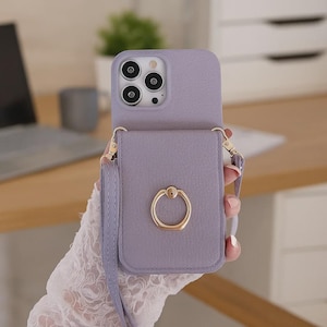 Peut inclure: Un étui de téléphone lavande avec une sangle assortie et un support de bague doré. L'étui a une finition texturée et est conçu pour contenir un téléphone. La sangle est fixée avec du matériel doré.