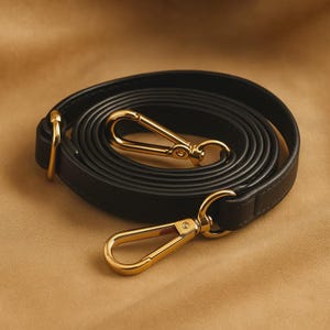 Puede incluir: Una correa de cuero negro enrollada con cierres y herrajes metálicos dorados. La correa está diseñada para un bolso o cartera, con una apariencia suave y elegante. Los herrajes dorados proporcionan un acento contrastante.