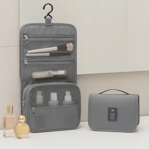 K&ouml;nnte beinhalten: Ein grauer Reise-Organizer zum Aufh&auml;ngen mit mehreren Rei&szlig;verschlussf&auml;chern und einem Haken zum Aufh&auml;ngen. Der Organizer ist ge&ouml;ffnet und zeigt Make-up-Pinsel und kleine Flaschen. Daneben liegt eine passende graue Reisetasche sowie zwei Parf&uuml;mflaschen.
