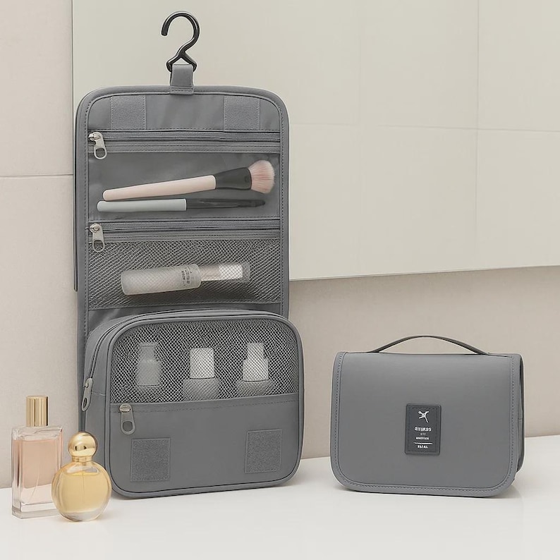 K&ouml;nnte beinhalten: Ein grauer Reise-Organizer zum Aufh&auml;ngen mit mehreren Rei&szlig;verschlussf&auml;chern und einem Haken zum Aufh&auml;ngen. Der Organizer ist ge&ouml;ffnet und zeigt Make-up-Pinsel und kleine Flaschen. Daneben liegt eine passende graue Reisetasche sowie zwei Parf&uuml;mflaschen.