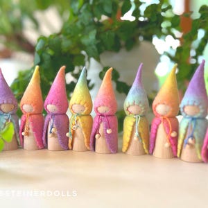 May include: Nine wooden peg dolls with rainbow-colored conical hats and capes. Each doll has a simple face and is arranged in a row. The text "DEBSSTEINERDOLLS" is visible at the bottom. The dolls are approximately 10 cm tall.