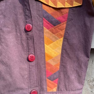 Może przedstawiać: Purpurowa kurtka dżinsowa z patchworkowym wzorem po prawej stronie. Patchwork wykonany jest z r&oacute;żnych odcieni pomarańczowego, czerwonego i ż&oacute;łtego materiału. Kurtka ma trzy czerwone guziki.