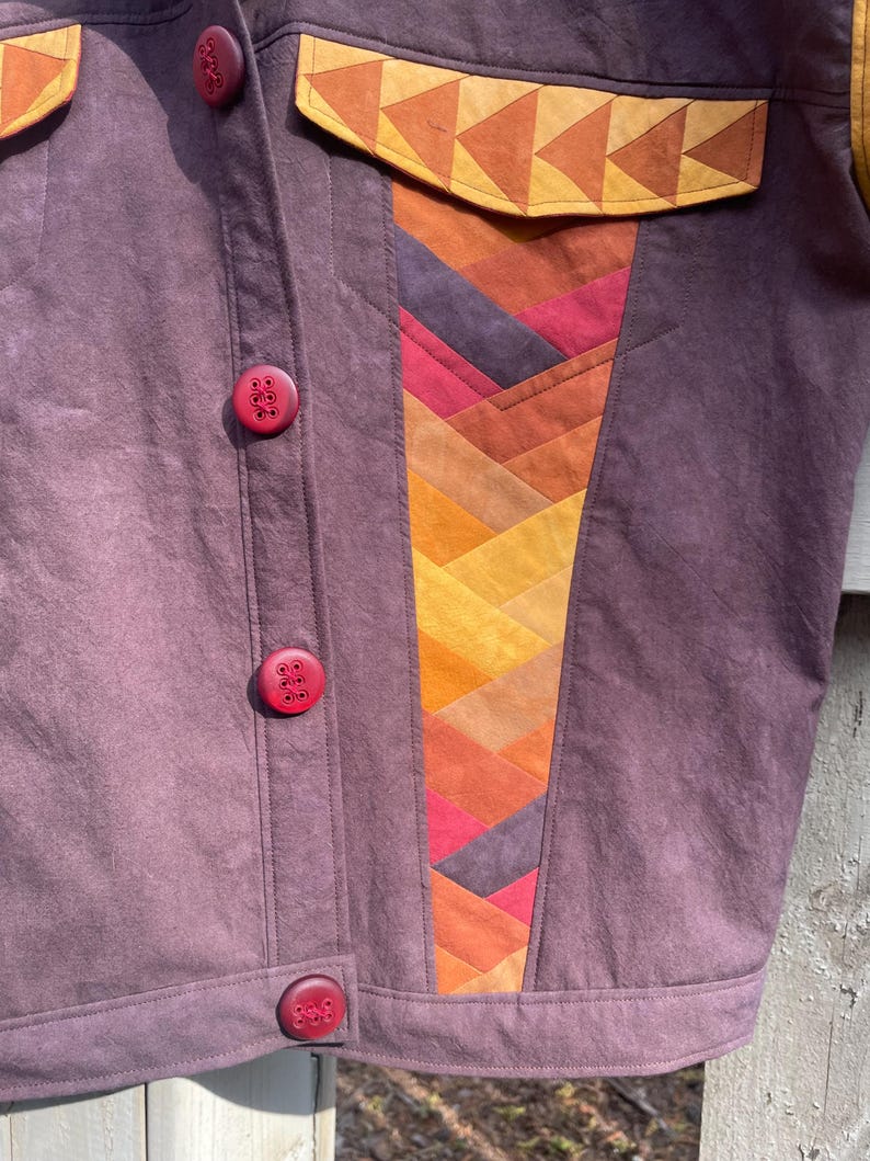 Może przedstawiać: Purpurowa kurtka dżinsowa z patchworkowym wzorem po prawej stronie. Patchwork wykonany jest z r&oacute;żnych odcieni pomarańczowego, czerwonego i ż&oacute;łtego materiału. Kurtka ma trzy czerwone guziki.