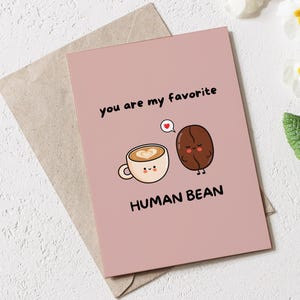 Puede incluir: Una tarjeta de felicitación rosa con el texto "you are my favorite HUMAN BEAN" e ilustraciones de una taza de café y un grano de café. La tarjeta está sobre un sobre marrón, con flores blancas y hojas verdes en el fondo.