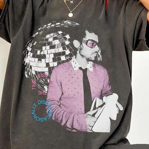 Puede incluir: Camiseta gris oscuro con un estampado gráfico de un hombre con gafas de sol y una bola de discoteca. El hombre lleva un suéter rosa y una corbata negra. El texto de la camiseta dice "Kiss the Disc occasionally."