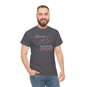 May include: A charcoal gray t-shirt featuring the text "Banda Karachi Ka" in a decorative script. The text is in red, black, and white. The shirt is a crew neck and is made of a soft material.