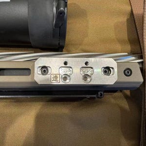 May include: Close-up of a rifle with a silver barrel and a tan-colored receiver. A silver metal plate with the Japanese character for "make" is attached to the receiver. The rifle is set against a tan background.