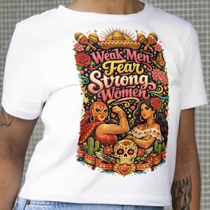 May include: White t-shirt featuring a colorful graphic with the text "Weak Men Fear Strong Women." The design includes illustrations of a luchadora, a woman with a rose, a sugar skull, and floral elements.